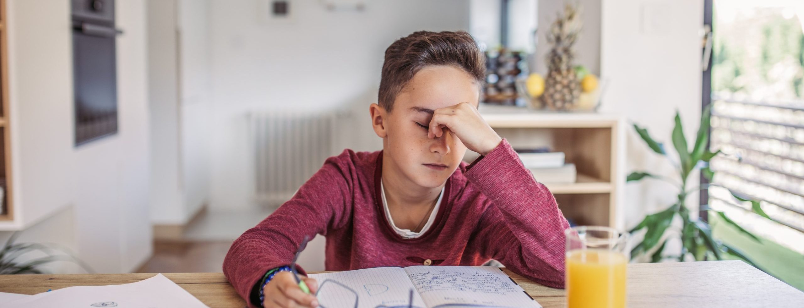 Alternate Text "A young boy sits at a wooden table with an open notebook, eyes closed and pinching the bridge of his nose in a gesture of frustration or exhaustion. He holds a pair of eyeglasses in his other hand, suggesting eye strain, while a glass of orange juice sits nearby.