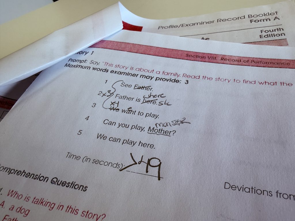 A close-up of a 'Profile/Examiner Record Booklet' showing a reading assessment for 'Story 1.' The simple text (e.g., 'See Father,' 'We want to play') is marked with handwritten examiner notations indicating reading errors, such as crossing out 'Father' and substituting 'here' with 'where.' The time is handwritten at the bottom as '>49' seconds.